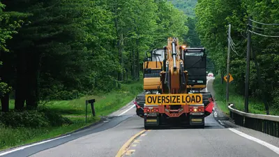 A flatbed truck hauling a large construction vehicle on a road. The truck has an orange oversized load banner on the back of it.