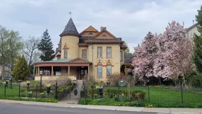 Beautiful old home with rounded covered porch and pointed peak, surrounded by green landscaping and a pink blossom tree.