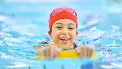 Child wearing a red swimming cap while swimming in a pool holding a yellow floating device.