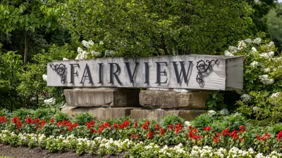 The Fairview Cemetery sign made on stone with black iron lettering. A flower bed and trees surround it.