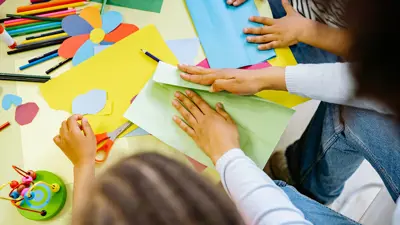 Craft supplies and coloured paper on a table with hands.