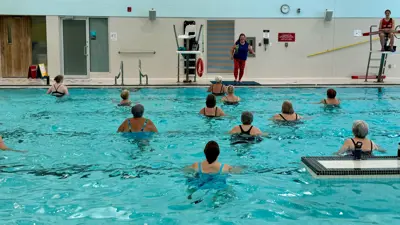 An aquafit class happening at MacBain Community Centre indoor pool. The instructor is on the pool deck and the participants are in the water facing the instructor. A lifeguard is elevated above, sitting in the lifeguard chair.