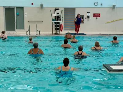 An aquafit class happening at MacBain Community Centre indoor pool. The instructor is on the pool deck and the participants are in the water facing the instructor. A lifeguard is elevated above, sitting in the lifeguard chair.