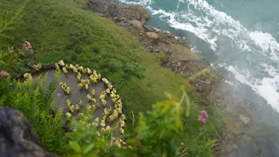 High-angle view of visitors in yellow rain ponchos gathered on an observation platform below Niagara Falls, with the rushing turquoise river, rocky shoreline, and lush green cliffs surrounding the scene.