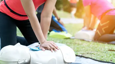 A close up photo of someone on their knees, doing chest compression on a white plastic dummy for a first aid or CPR class.