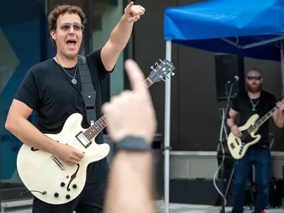 A guitarist performs on an outdoor stage as audience members watch and cheer during a live music performance.