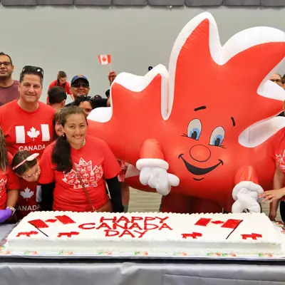 A group of people with a mascot dressed as a red maple leaf, posing behind the Happy Canada Day cake.
