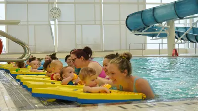 Caregivers with their babies in the MacBain Community Center indoor pool. The babies are sitting in yellow floatation devices, all in a row with their caregiver in front of them.