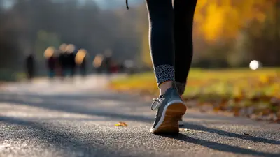 Image of a person walking on a paved trail, close up of the bottom portion of their legs and fee. The background has blurry images of people walking ahead. The trees have autumn coloured leaves